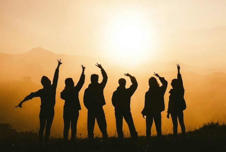 Teamarbeit Sechs Personen in Silhouette heben die Arme vor einem Sonnenuntergang.